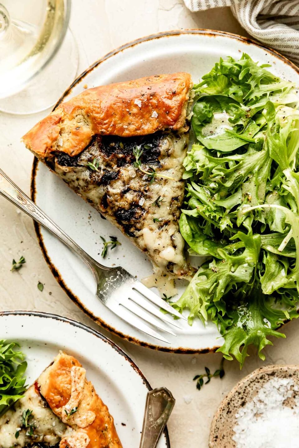 An overhead shot of a slice of a cheesy caramelized onion galette alongside mixed greens on a white plate atop an off-white surface. A glass of white wine sits alongside the plate.
