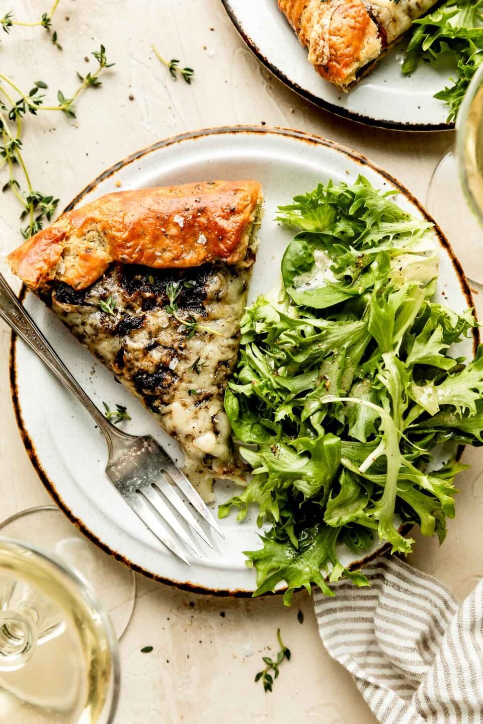 An overhead shot of a slice of a caramelized onion galette alongside mixed greens on a white plate atop an off-white surface. Two glasses of white wine sit alongside the plate.