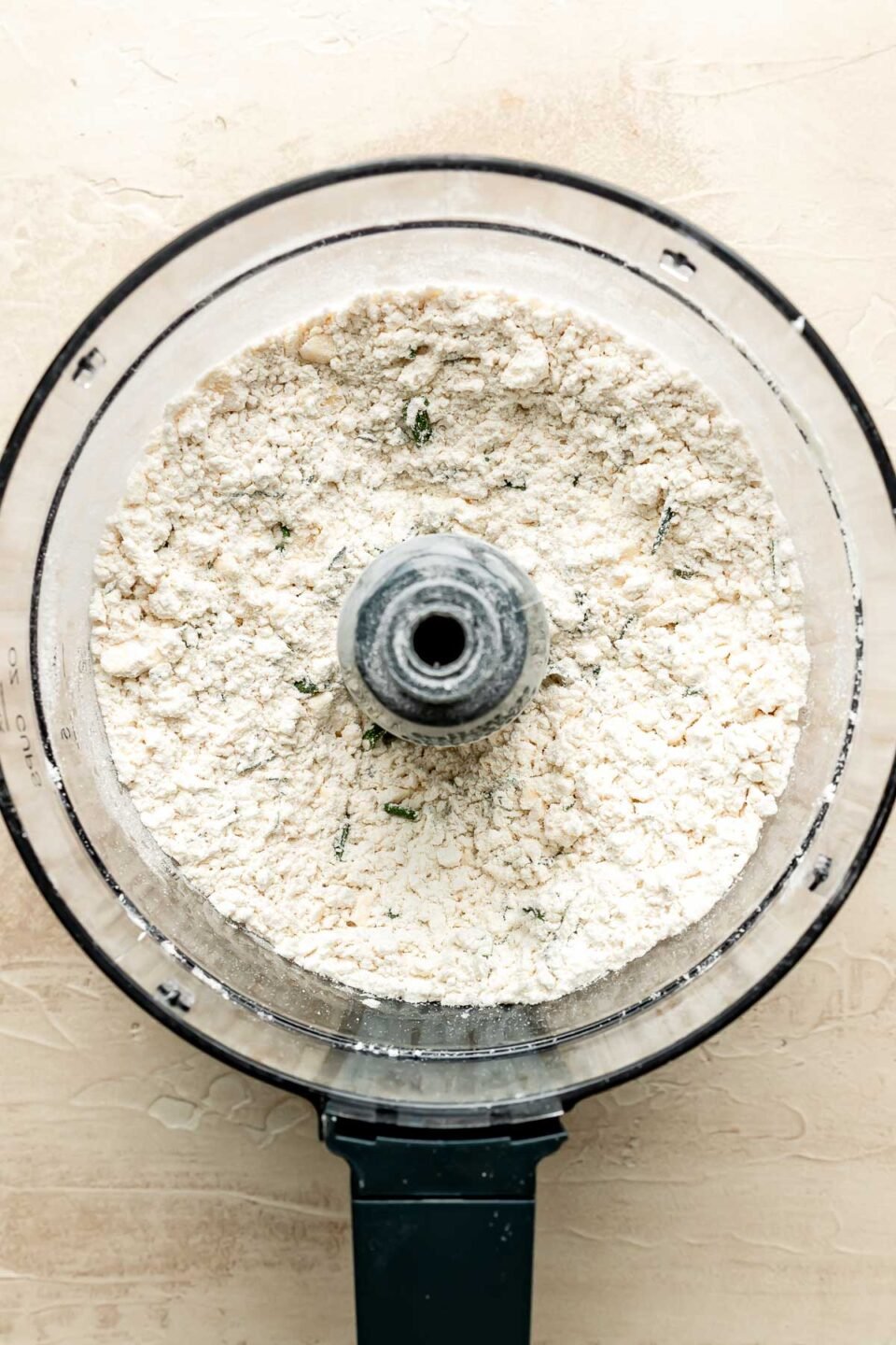 An overhead shot of flour and chopped herbs in the bowl of a food processor atop an off-white surface.
