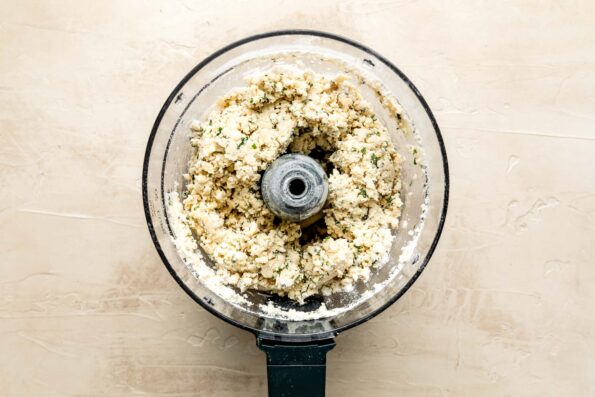 An overhead shot of a flour and butter crust mixture in the bowl of a food processor atop an off-white surface.
