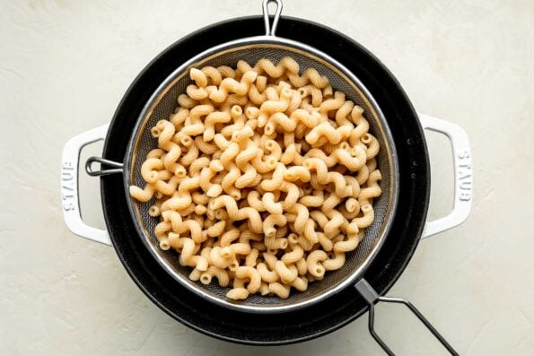 An overhead shot of cooked cavateppi pasta in a colander over a cast iron cocotte on an off-white surface.