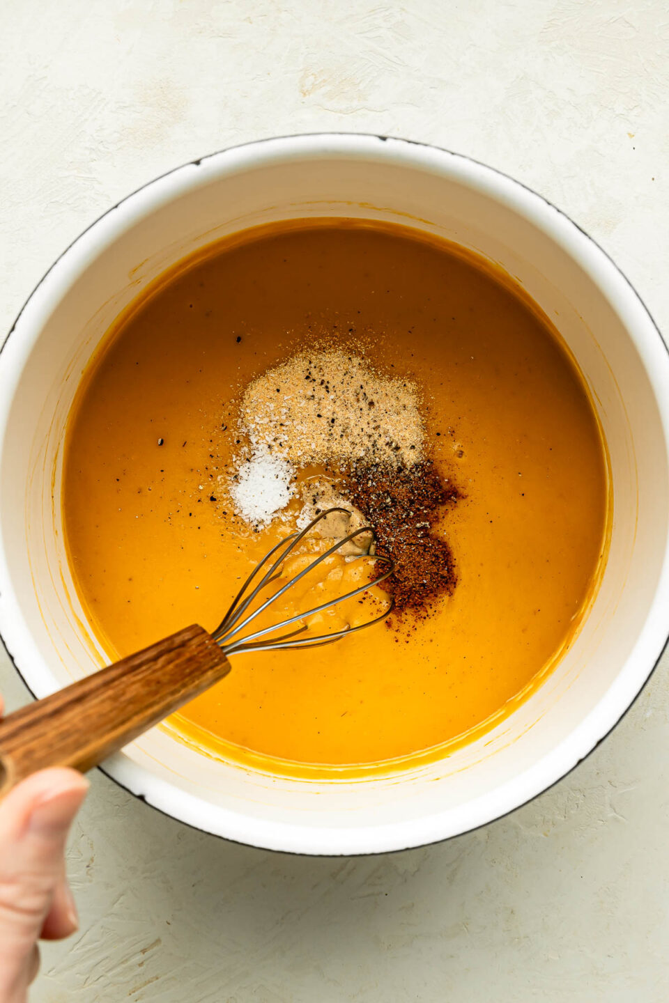 An overhead shot of a woman's hand whisking a saucepan of soup and dried spices on an off-white surface.