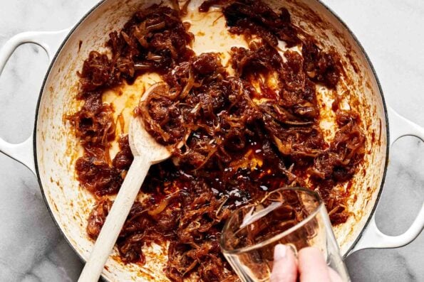 An overhead shot of a woman's hand pouring sherry into dark brown caramelized onions in a white skillet atop a white marbled surface.