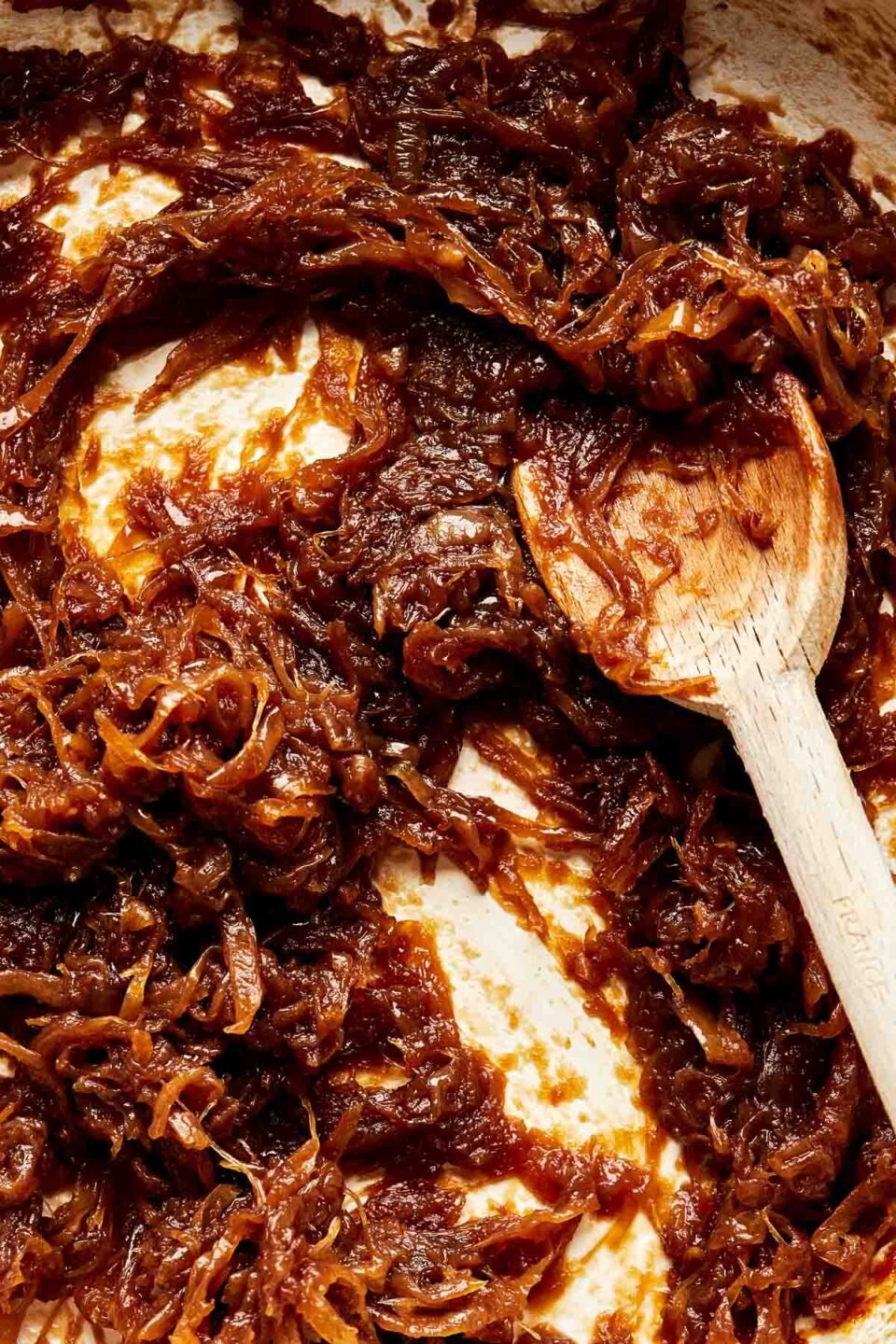 A close-up overhead shot of a wooden spoon stirring caramelized onions in a white skillet.