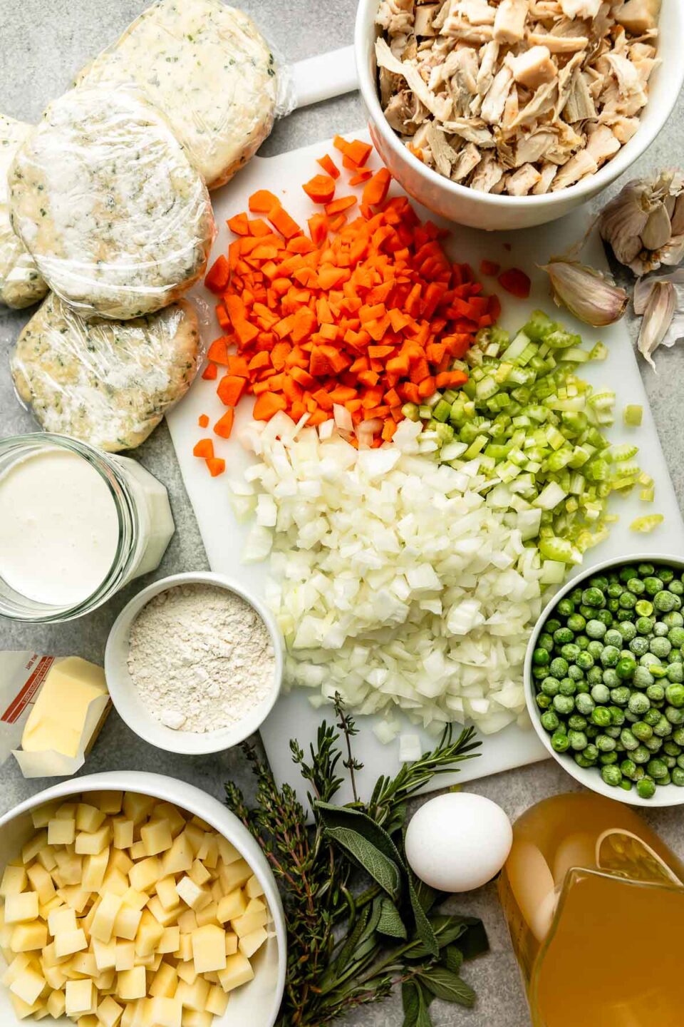 An overhead shot of ingredients displayed on a white cutting board and various bowls on a grey surface: chopped carrots, celery, onion, and potato, fresh herbs, frozen peas, shredded chicken, an egg, individual crusts wrapped in plastic wrap, garlic, butter, flour, chicken stock, and heavy cream.