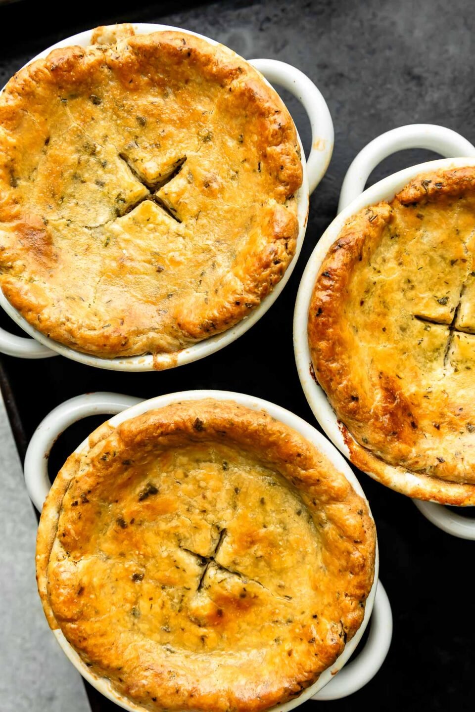 An overhead shot of three baked individual pot pies on a black tray atop a grey surface.