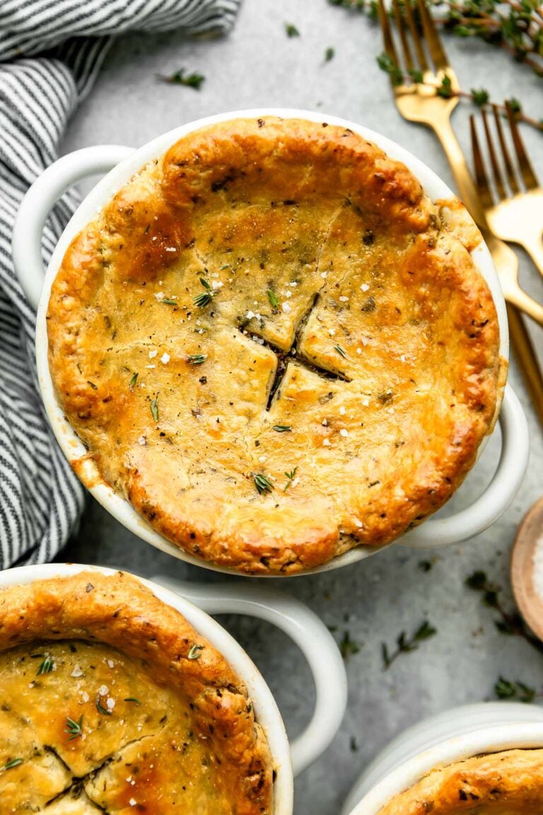 An overhead shot of an individual pot pie in a white ramekin atop a grey surface. A striped cloth, two gold forks and two additional pot pies sit alongside it.