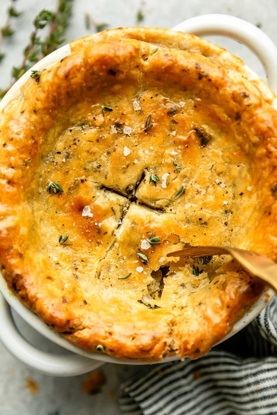 A close-up overhead shot of a fork digging into an individual pot pie on a grey surface alongside a striped cloth and fresh herbs.