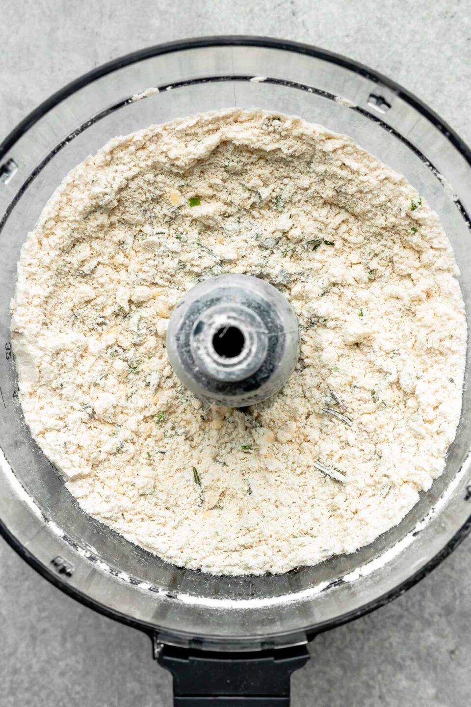 An overhead shot of dry pie crust ingredients in a food processor atop a grey textured surface.