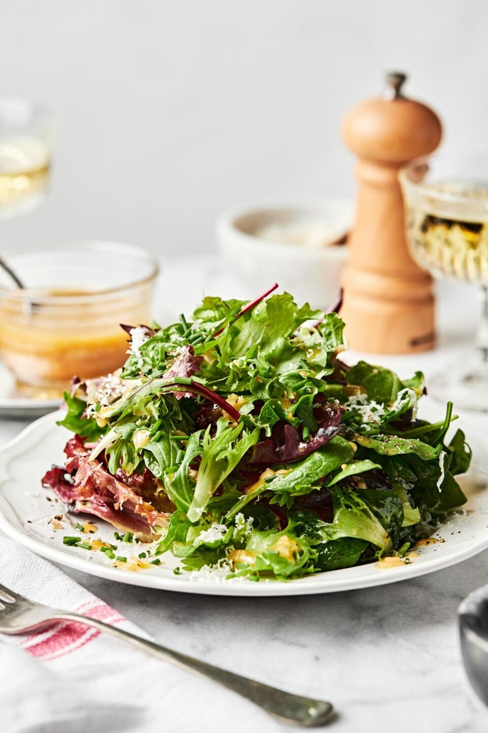 A side shot of bistro greens piled high on a white plate atop a white marbled surface. In the background, there is a glass bowl of dressing, two glasses of wine and a pepper grinder.
