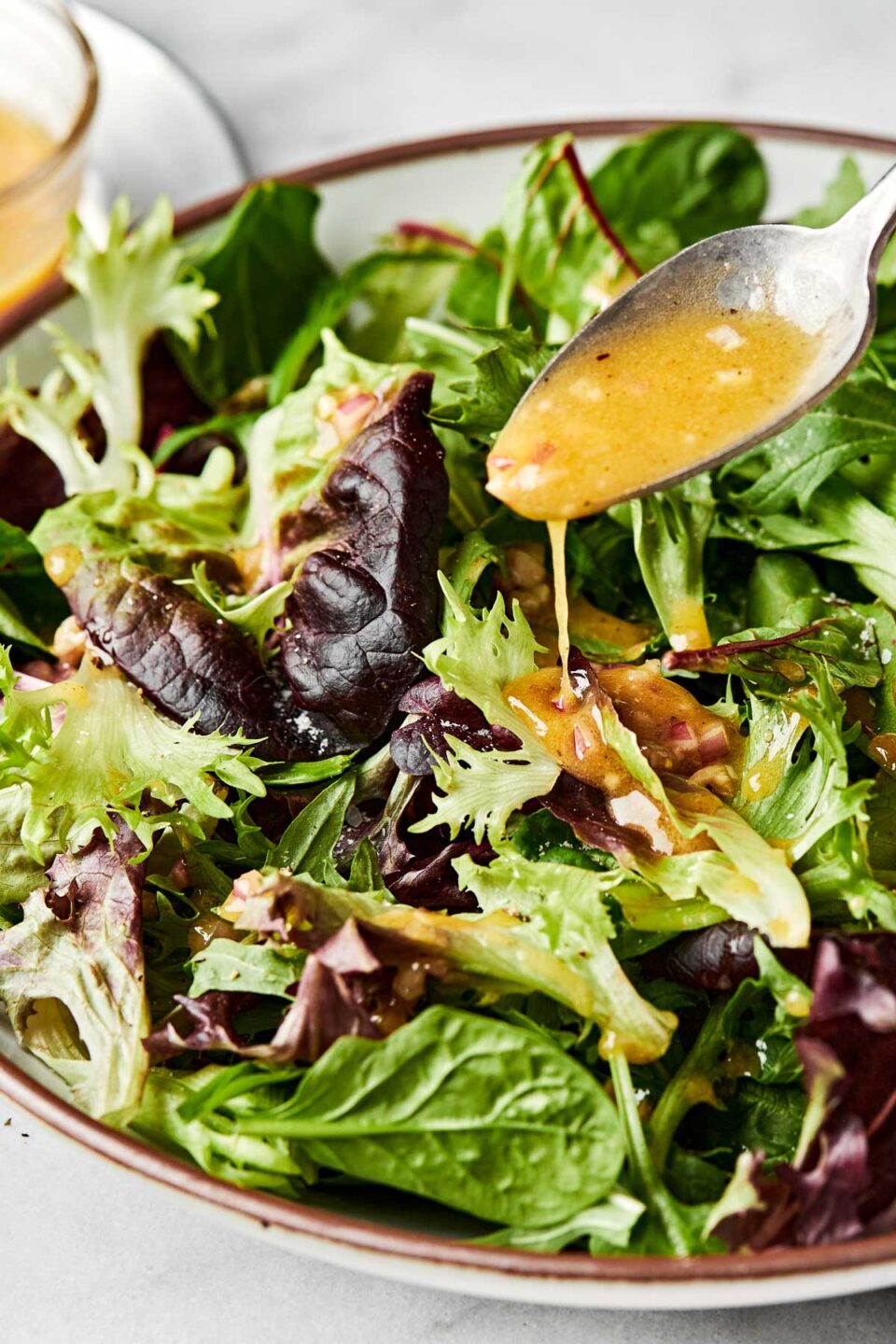 A close-up shot of a spoon drizzling dressing over a bowl of mixed greens.