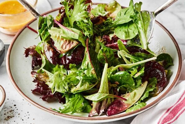 A close up shot of dressed mixed greens being tossed with two large spoons in a shallow bowl. A small bowl of dressing sits in the background.
