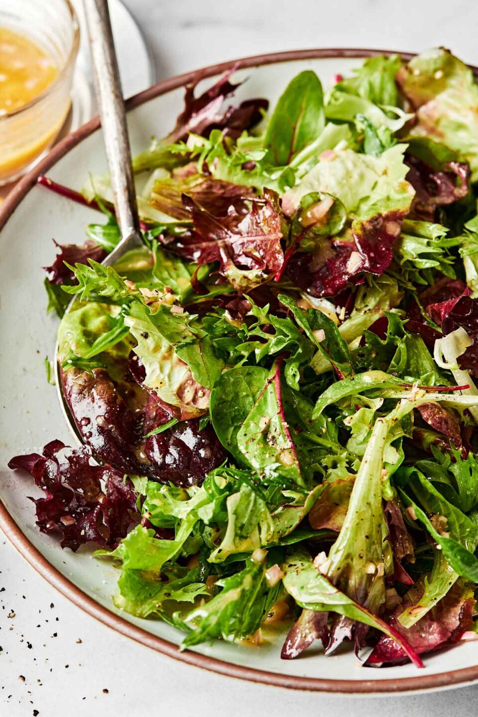 A close up shot of dressed mixed greens in a shallow bowl on a white surface.