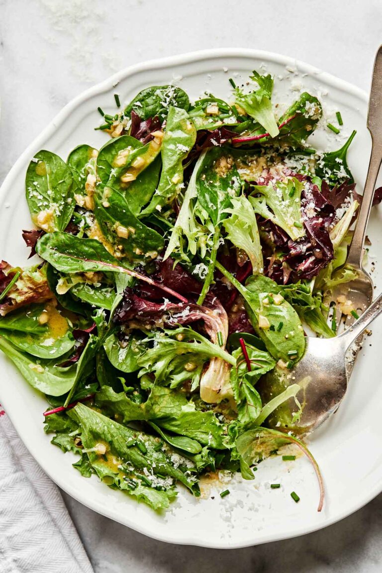 An overhead shot of a mixed green side salad on a white scalloped plate atop a white surface.