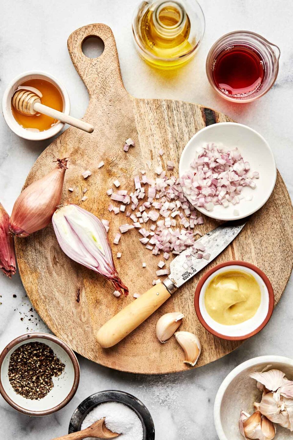 An overhead shot of ingredients displayed in bowls and on a wooden board atop a white marbled surface: halved and diced shallots, red wine vinegar, dijon mustard, salt, pepper, honey, olive oil, and garlic.