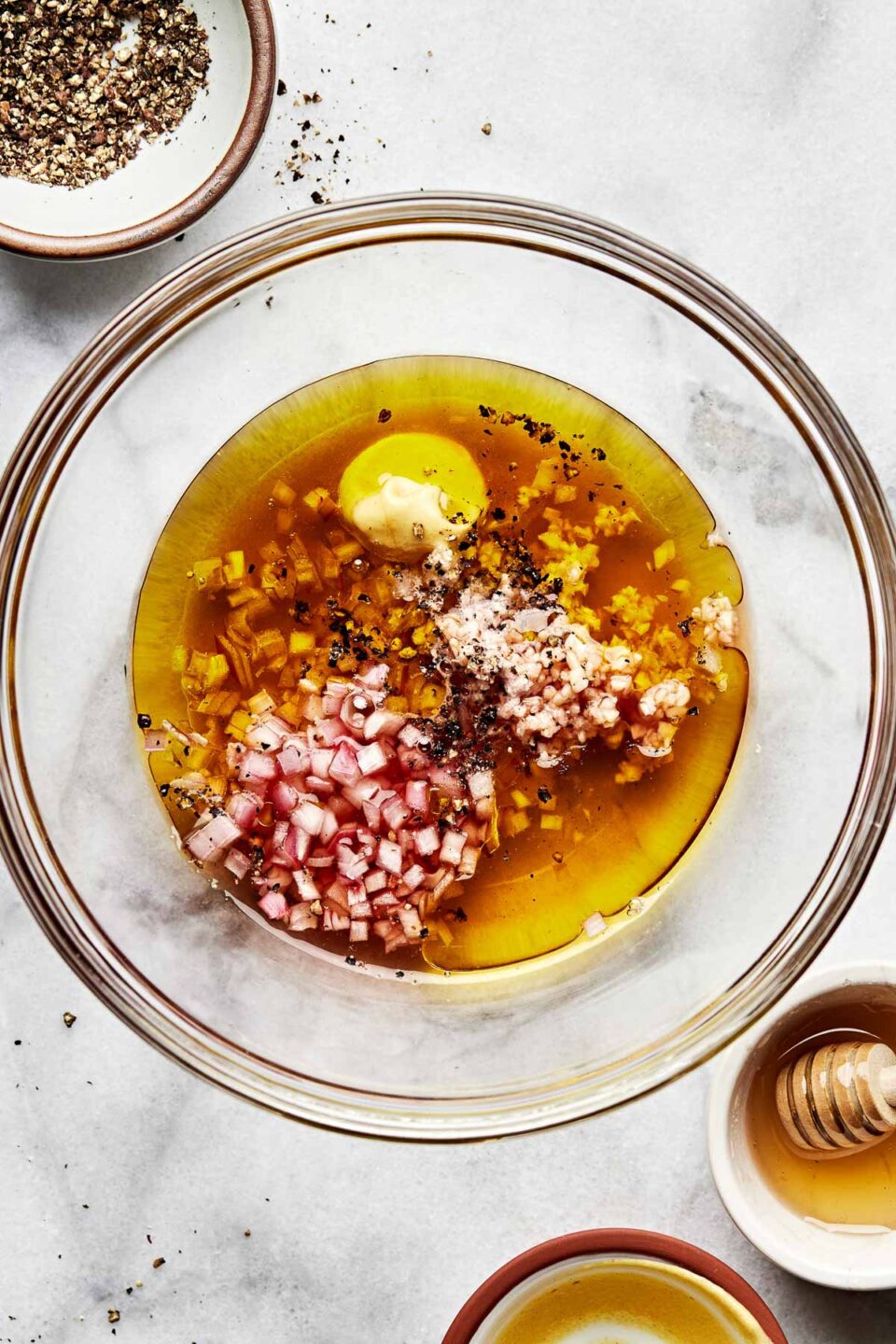 An overhead shot of shallots, olive oil, red wine vinegar, garlic, salt, pepper, honey and dijon mustard in a large glass bowl atop a white surface. Small dishes of pepper and honey sit alongside the bowl.