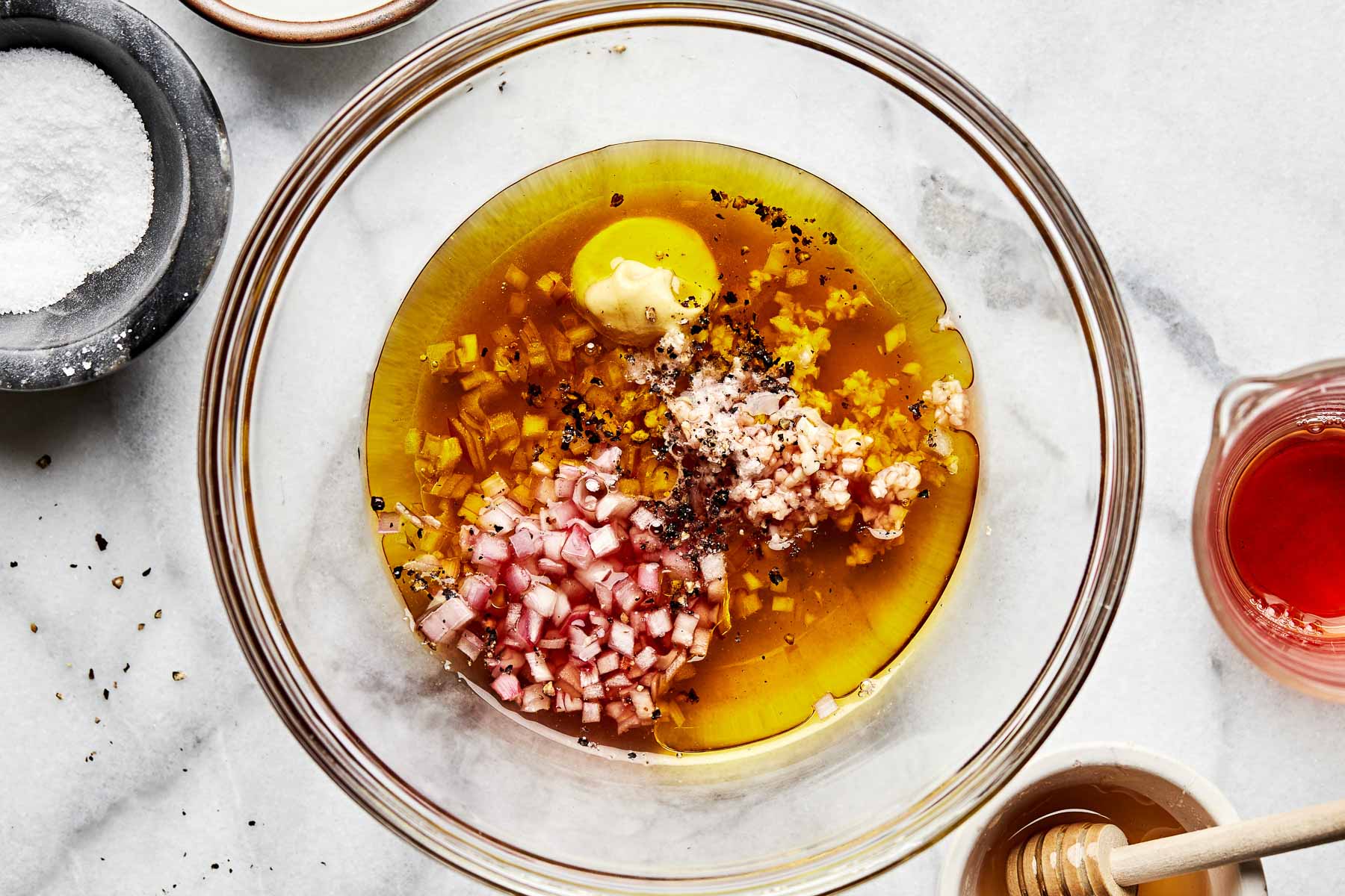 An overhead shot of shallots, olive oil, red wine vinegar, garlic, salt, pepper, honey and dijon mustard in a large glass bowl atop a white surface. Small dishes of salt, vinegar and honey sit alongside the bowl.