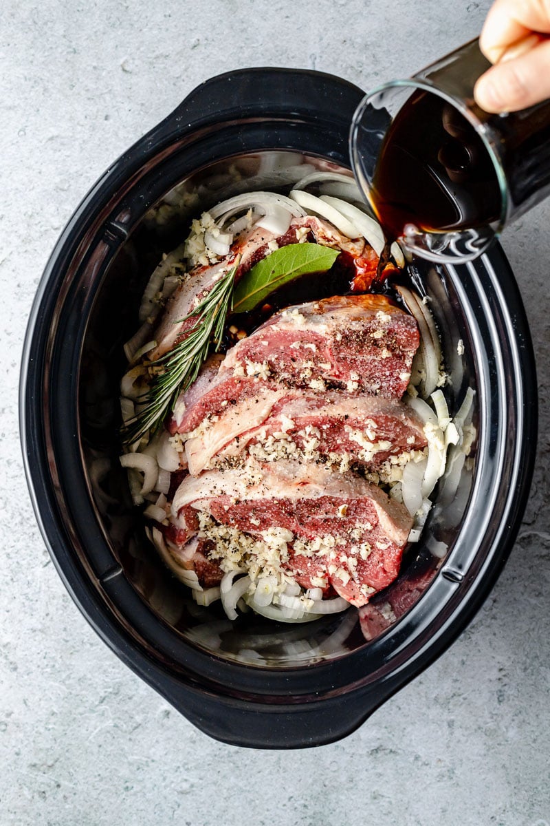 Lamb shoulder, sliced onion, garlic, rosemary, bay leaf, kosher salt, and ground black pepper are placed inside a slow cooker. A woman's hand holds a glass measuring cup and pours in a braising liquid made with aged balsamic vinegar, beef stock, and pure maple syrup over the prepared lamb.