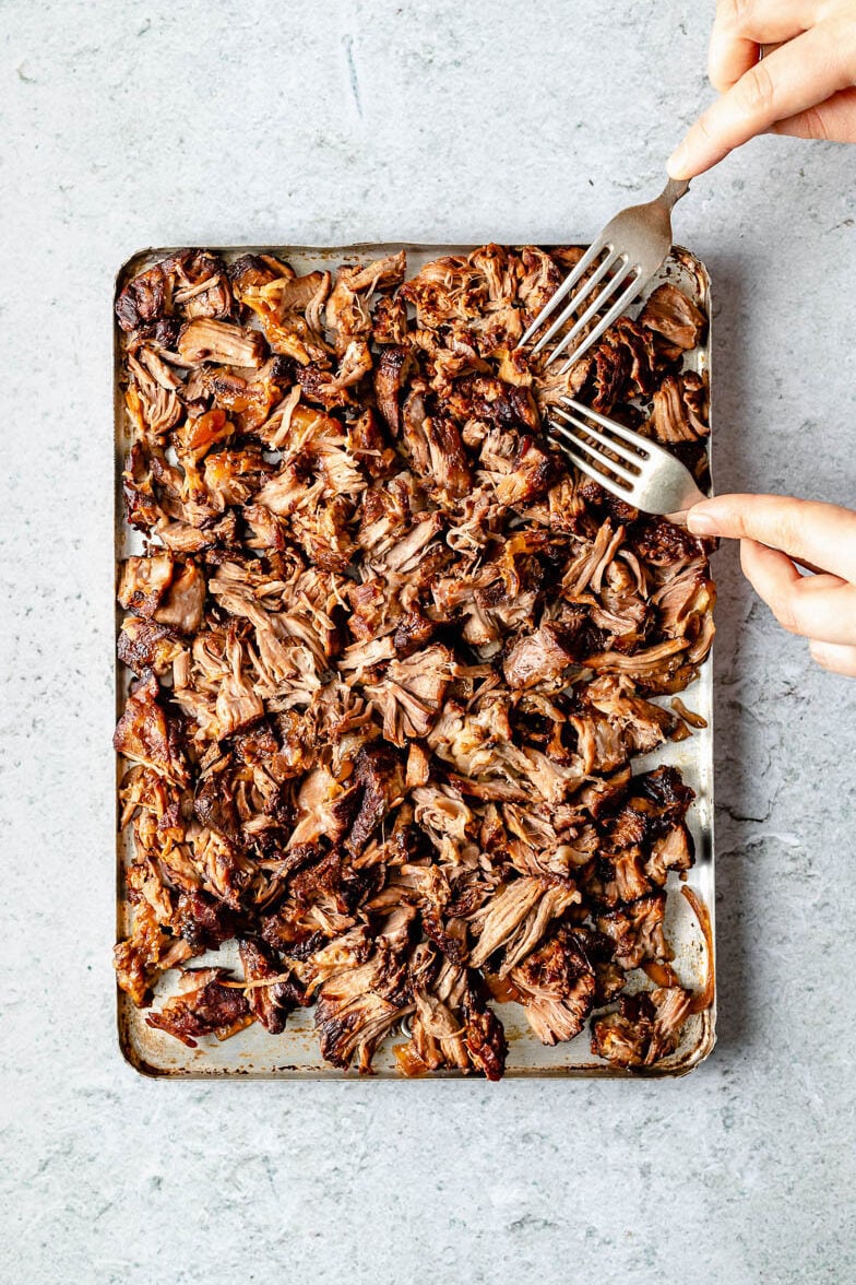 Shredded lamb fills an aluminum baking sheet. A woman's hands hold two forks and uses them to shred the lamb. The baking sheet sits atop a grey textured surface.