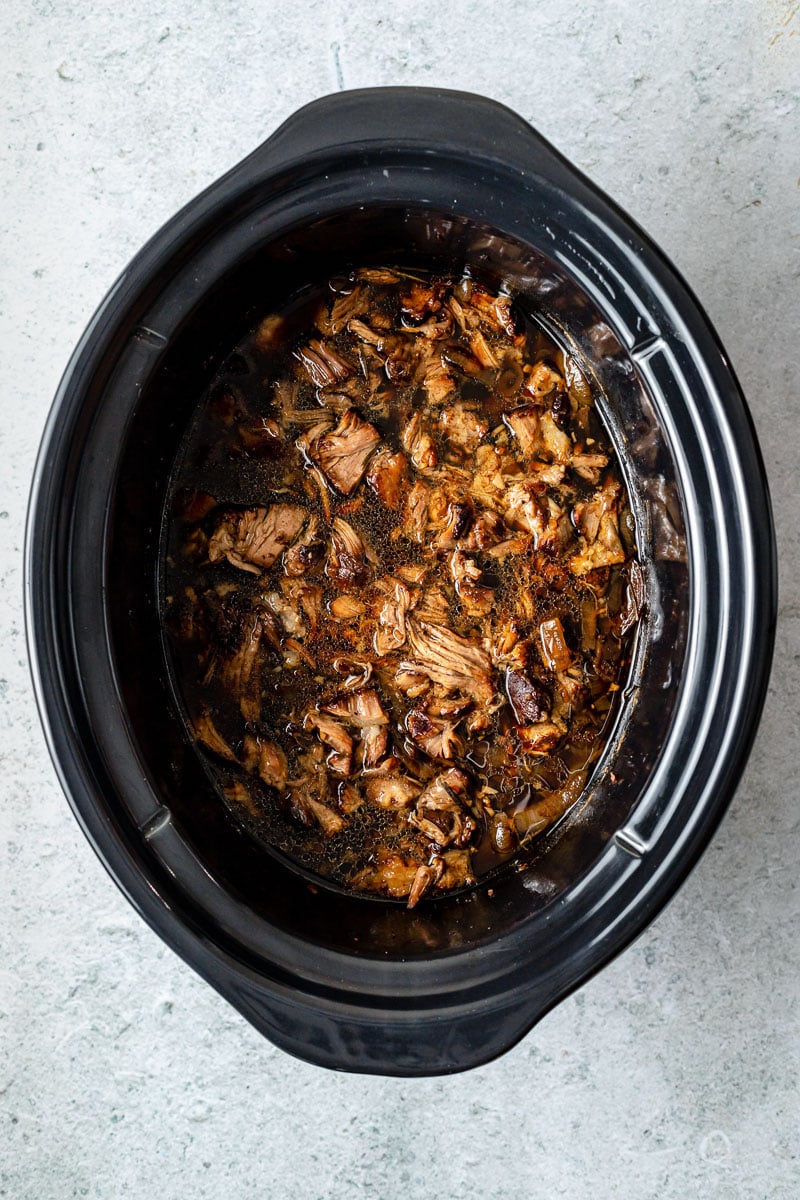 Shredded lamb fills a slow cooker in braising liquid. The slow cooker sits atop a grey textured surface.