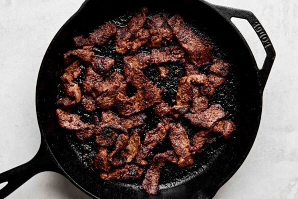 An overhead shot of seared beef in a black cast iron skillet atop a white surface.