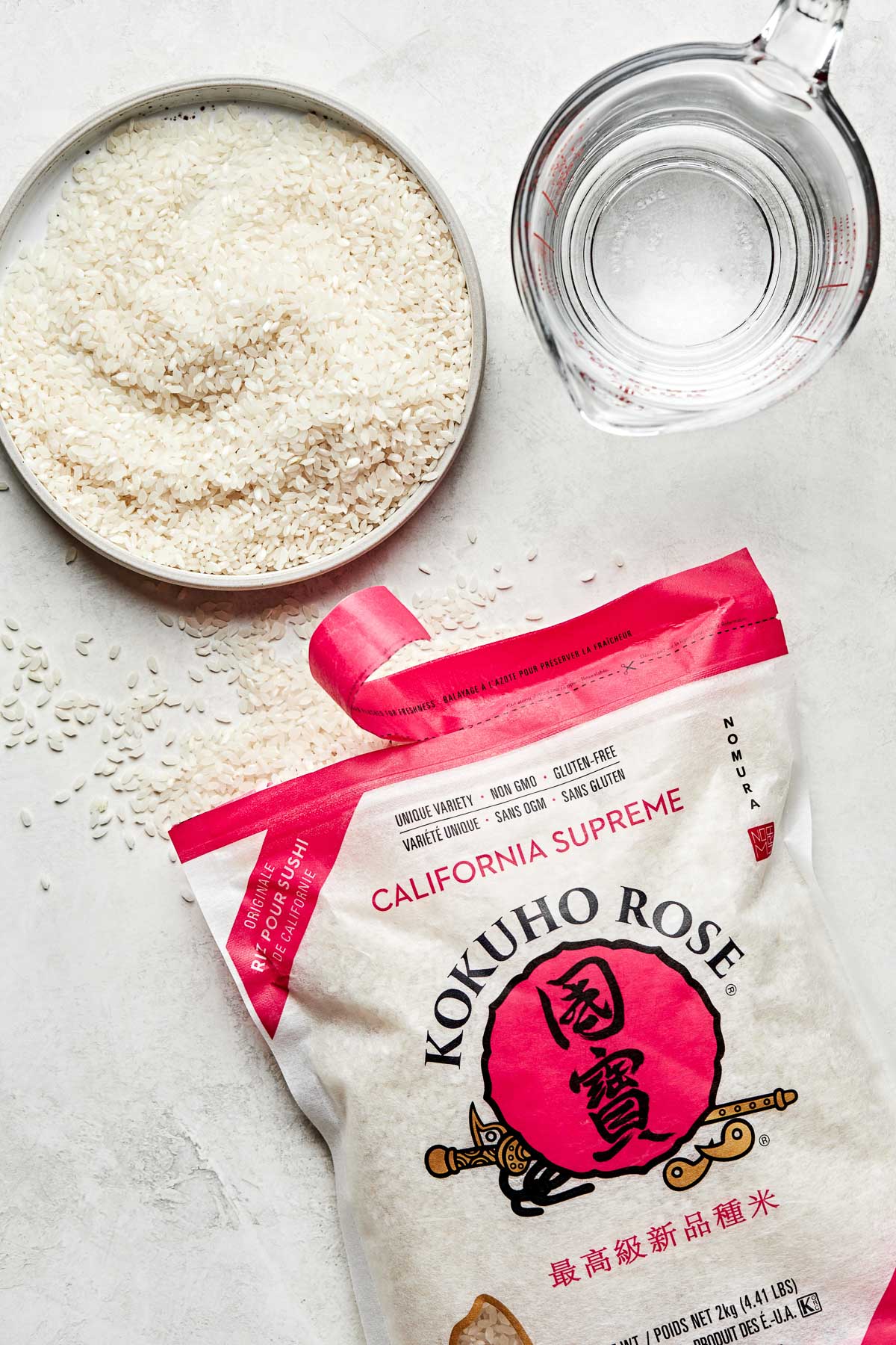 An overhead shot of a bag of uncooked calrose rice, some of which that has been poured onto a small white plate. A glass measuring cup of water sits alongside it on a white surface.