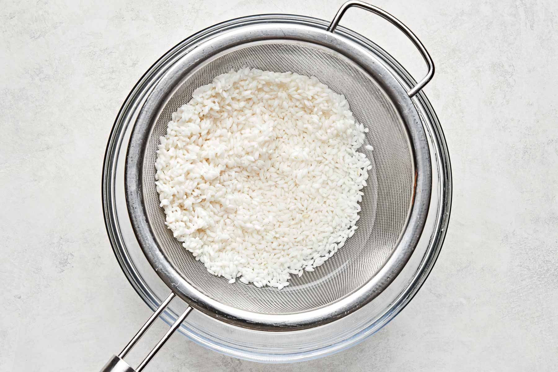 An overhead shot of rinsed rice being strained with a colander over a glass bowl atop a white surface.