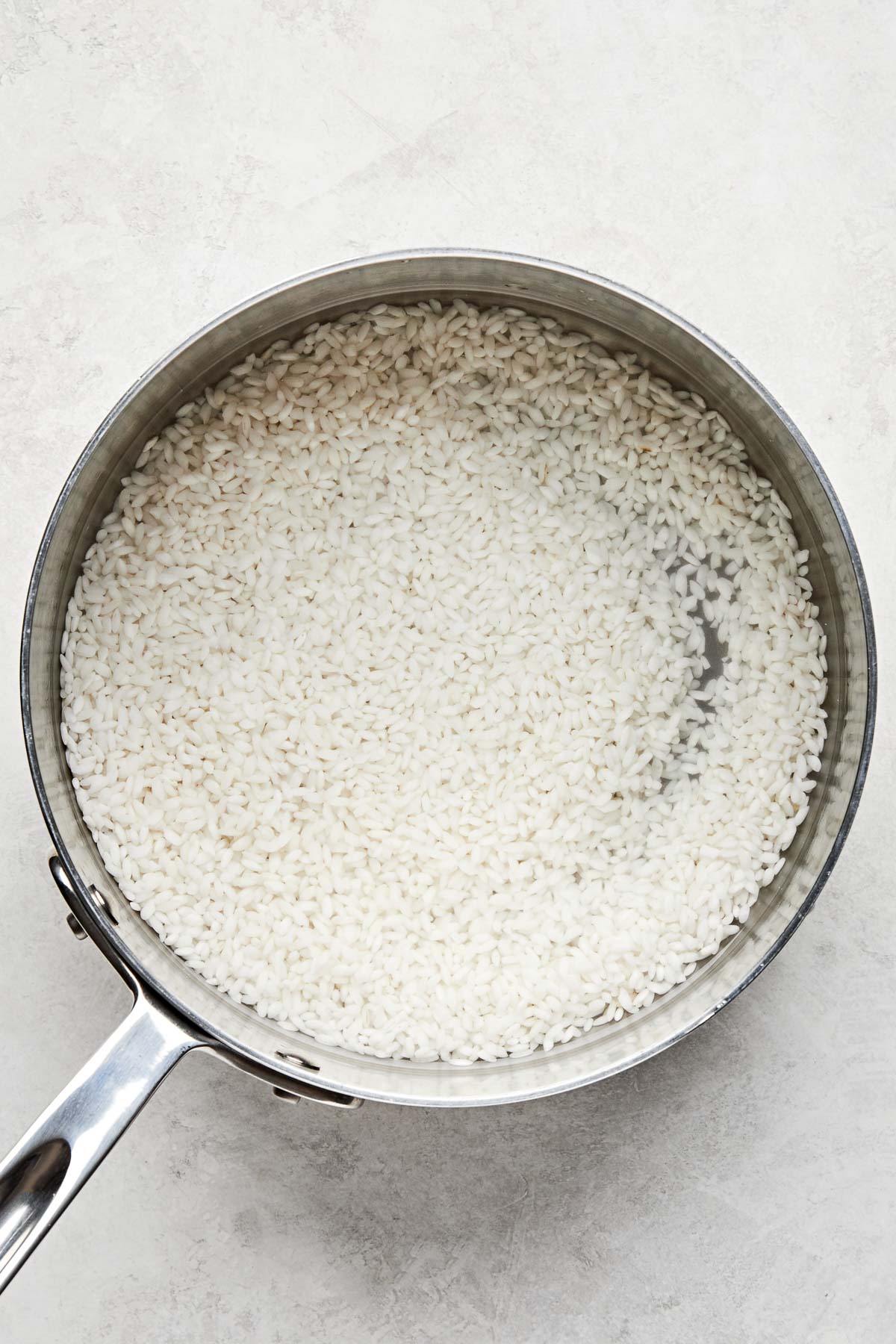 An overhead shot of rinsed, uncooked rice with water in a stainless steel pot atop a white marbled surface.