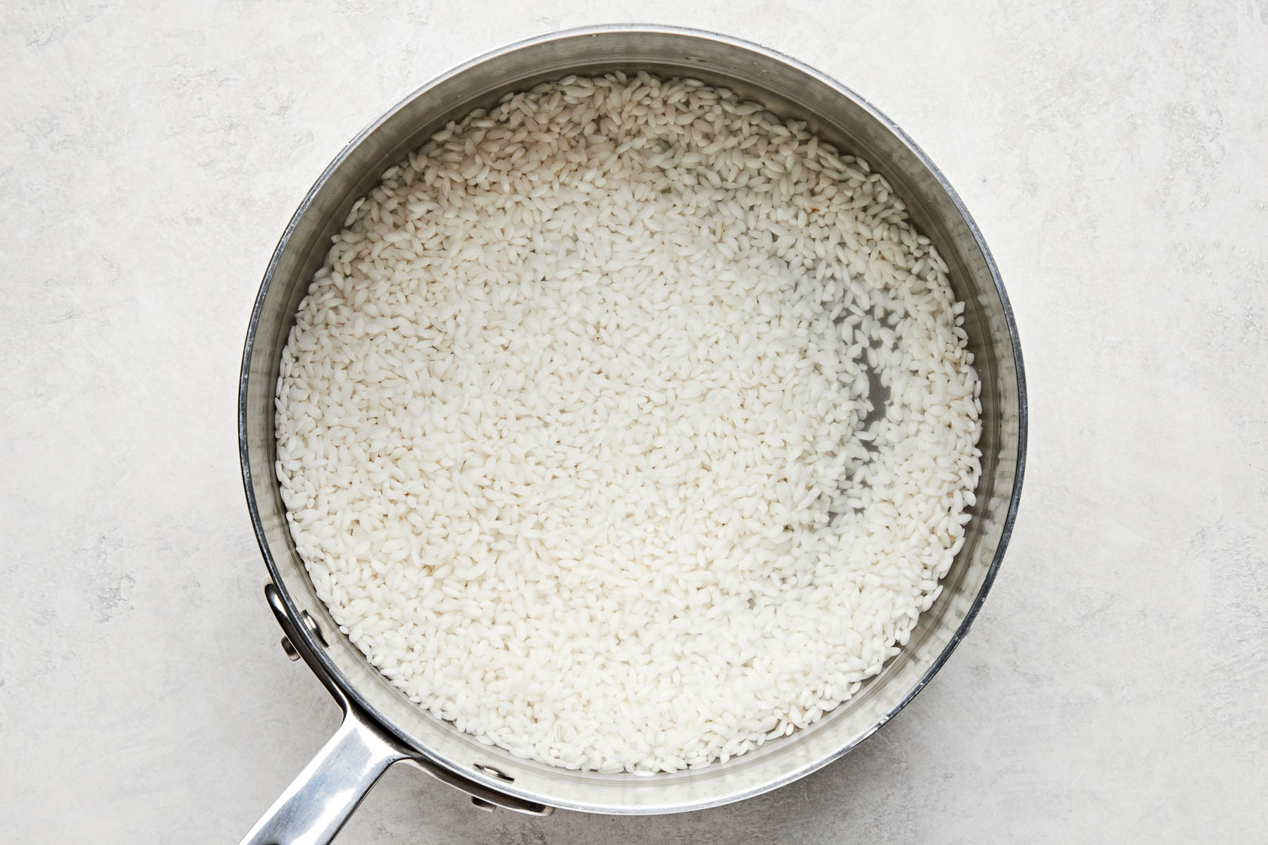 An overhead shot of rinsed, uncooked rice with water in a stainless steel pot atop a white marbled surface.