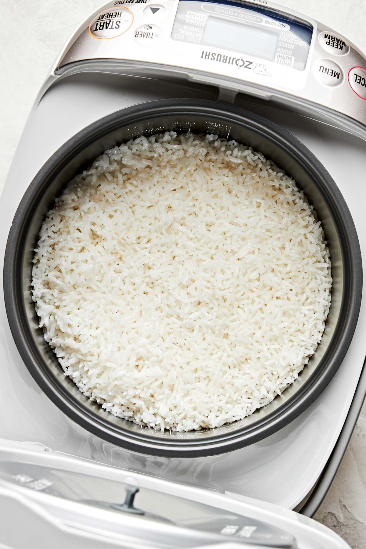 An overhead shot of cooked white rice in a rice cooker atop a white surface.