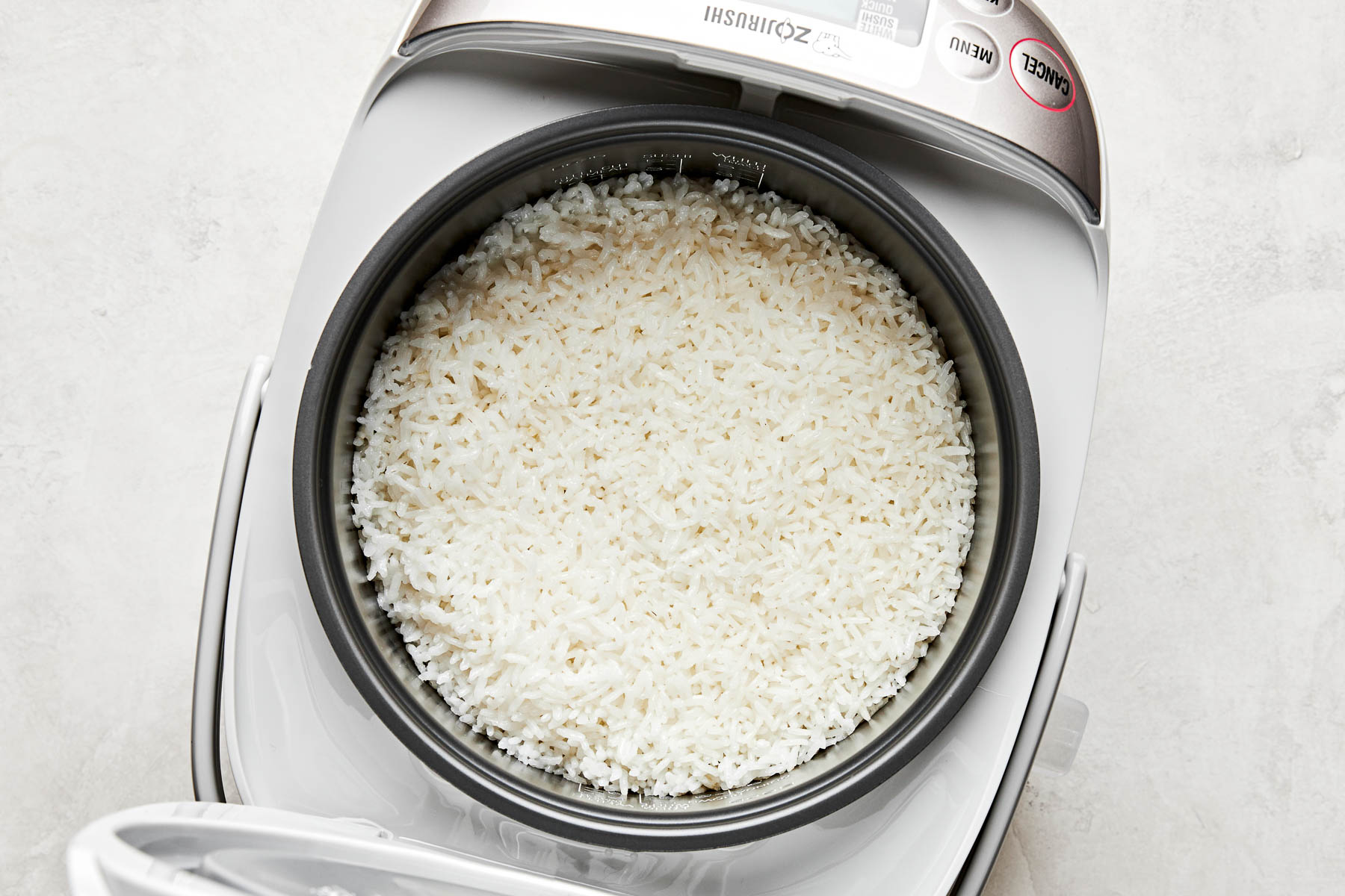 An overhead shot of cooked Calrose rice in a rice cooker atop a white surface.