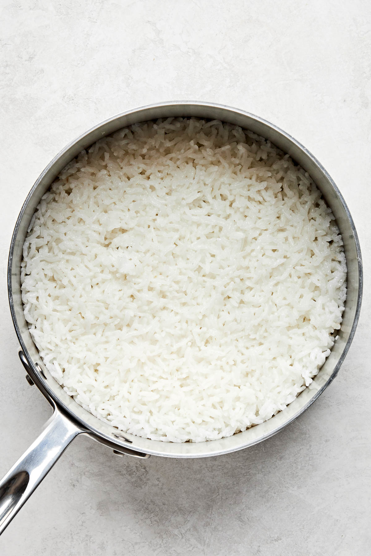An overhead shot of cooked Calrose rice in a stainless steel saucepan atop a white surface.
