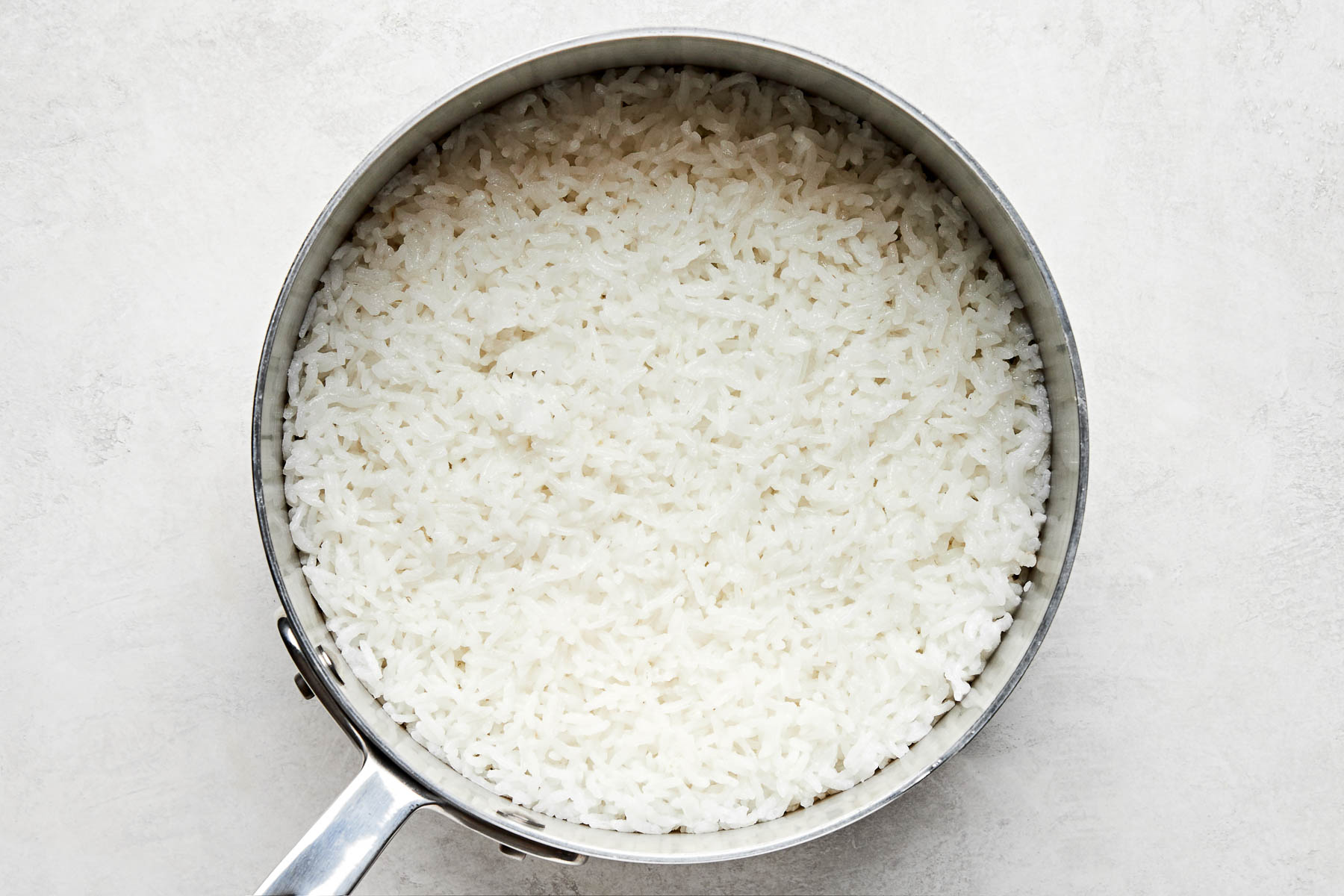 An overhead shot of cooked Calrose rice in a stainless steel saucepan atop a white surface.