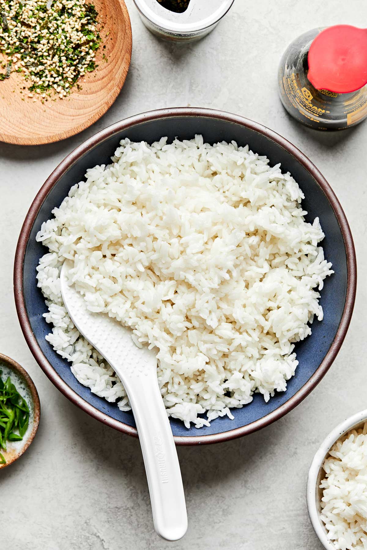 An overhead shot of fluffy cooked Calrose rice with a rice paddle in a blue bowl atop a light grey surface. Bowls of furikake, green onions and more cooked rice sit alongside it.