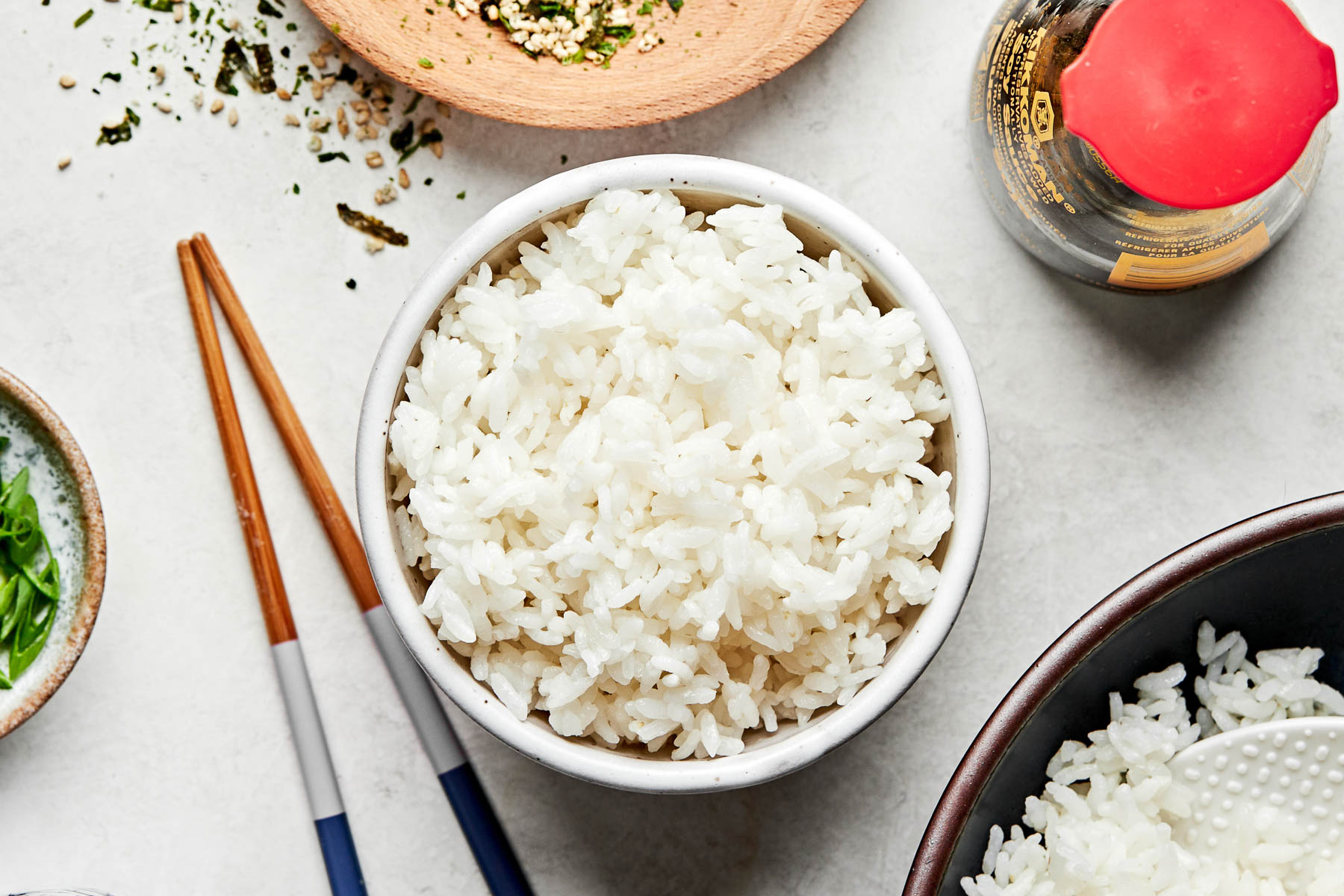 An overhead shot of cooked white rice in a small bowl atop a light grey surface. Chopsticks, a jar of soy sauce, a larger bowl of rice, and small bowls of furikake and green onions sit alongside it.