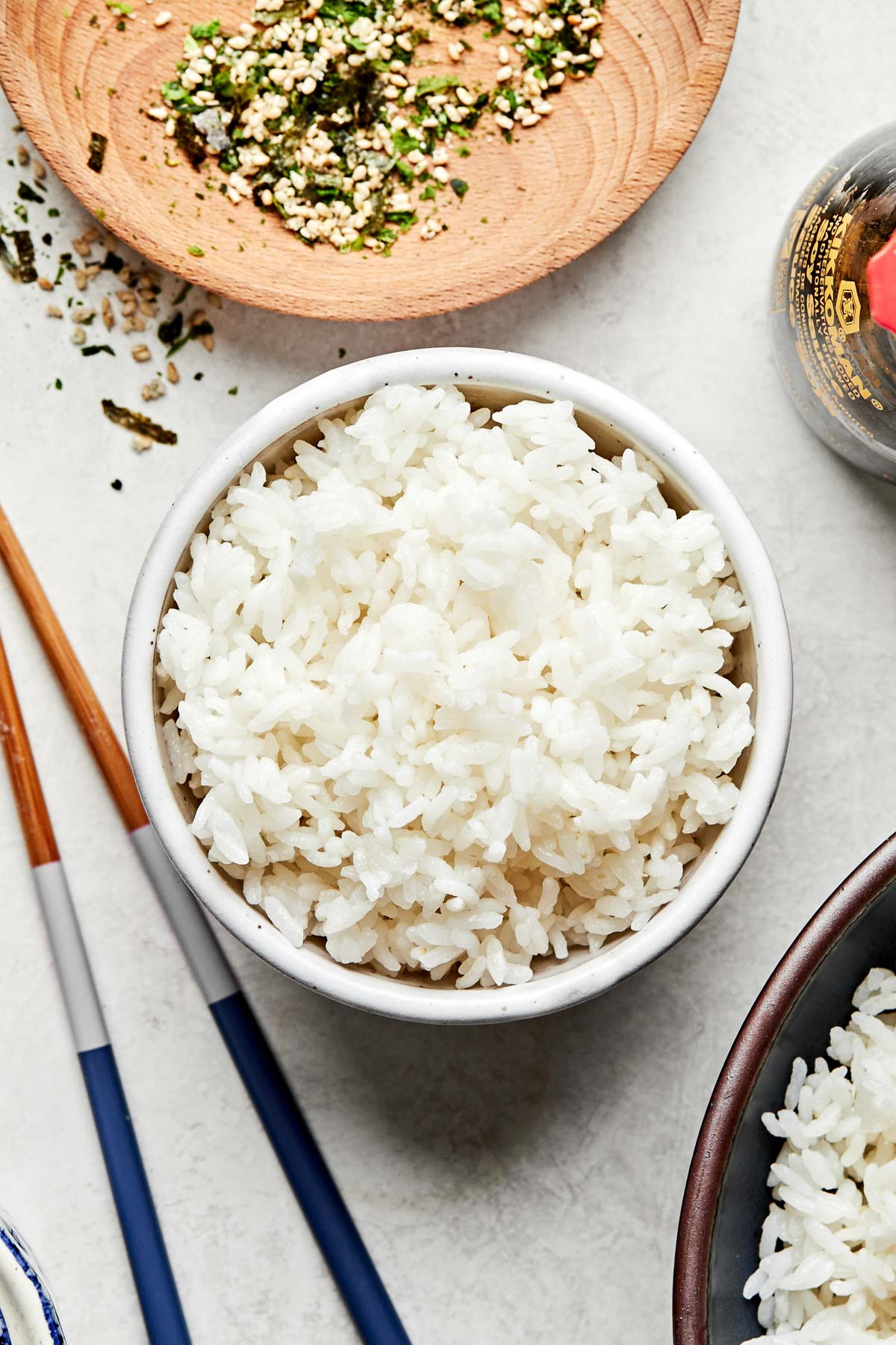 An overhead shot of cooked calrose rice in a small bowl atop a light grey surface. Chopsticks, a jar of soy sauce, a larger bowl of rice, and small bowls of furikake and green onions sit alongside it.