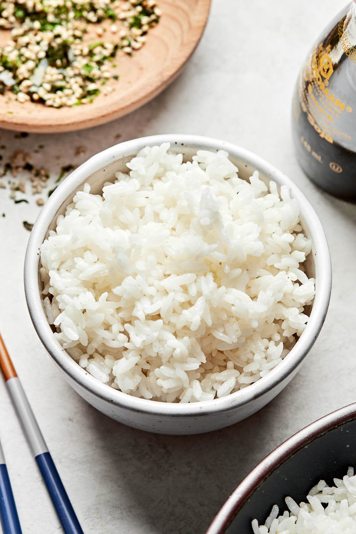 An angled overhead shot of fluffy calrose rice in a white bowl atop a light grey surface, alongside a jar of soy sauce and bowls of additional rice and furikake.