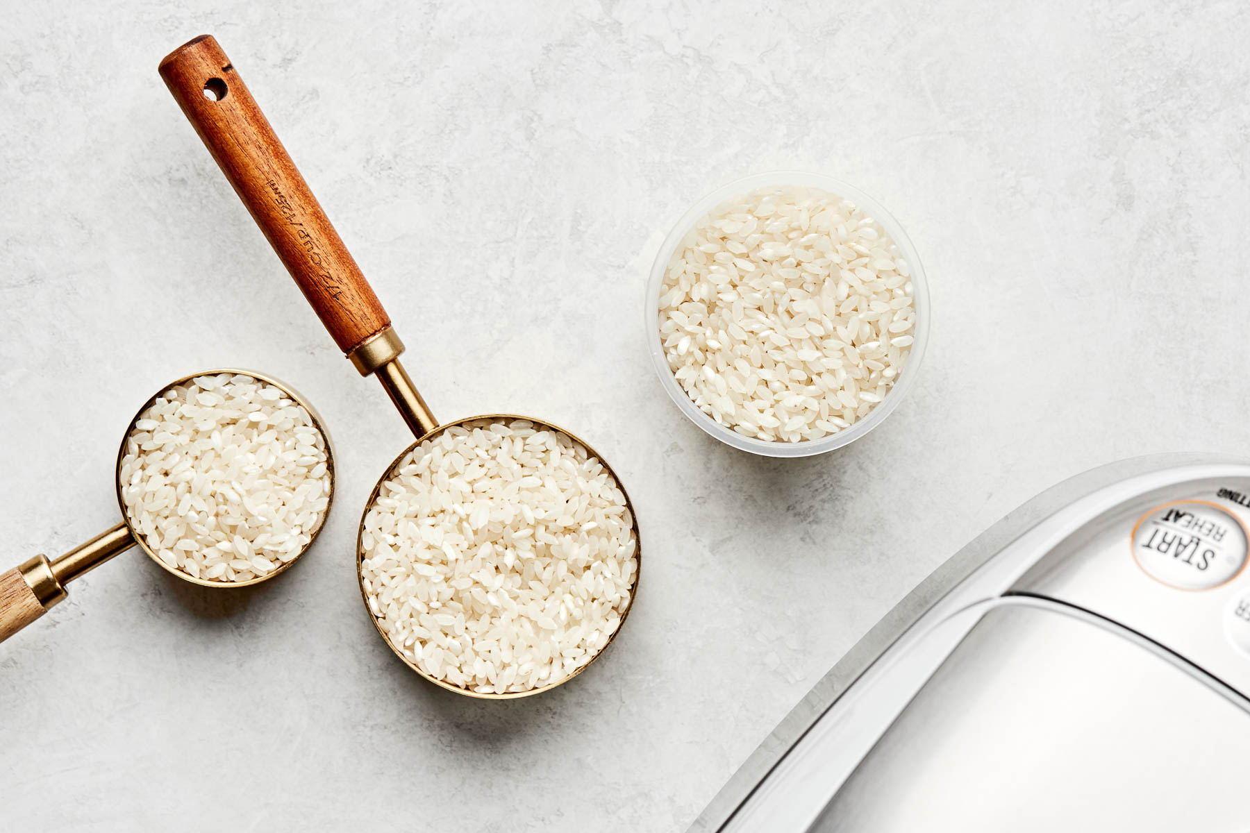 An overhead shot of three different measuring cups with uncooked calrose rice on a white marbled surface alongside the edge of a rice cooker.