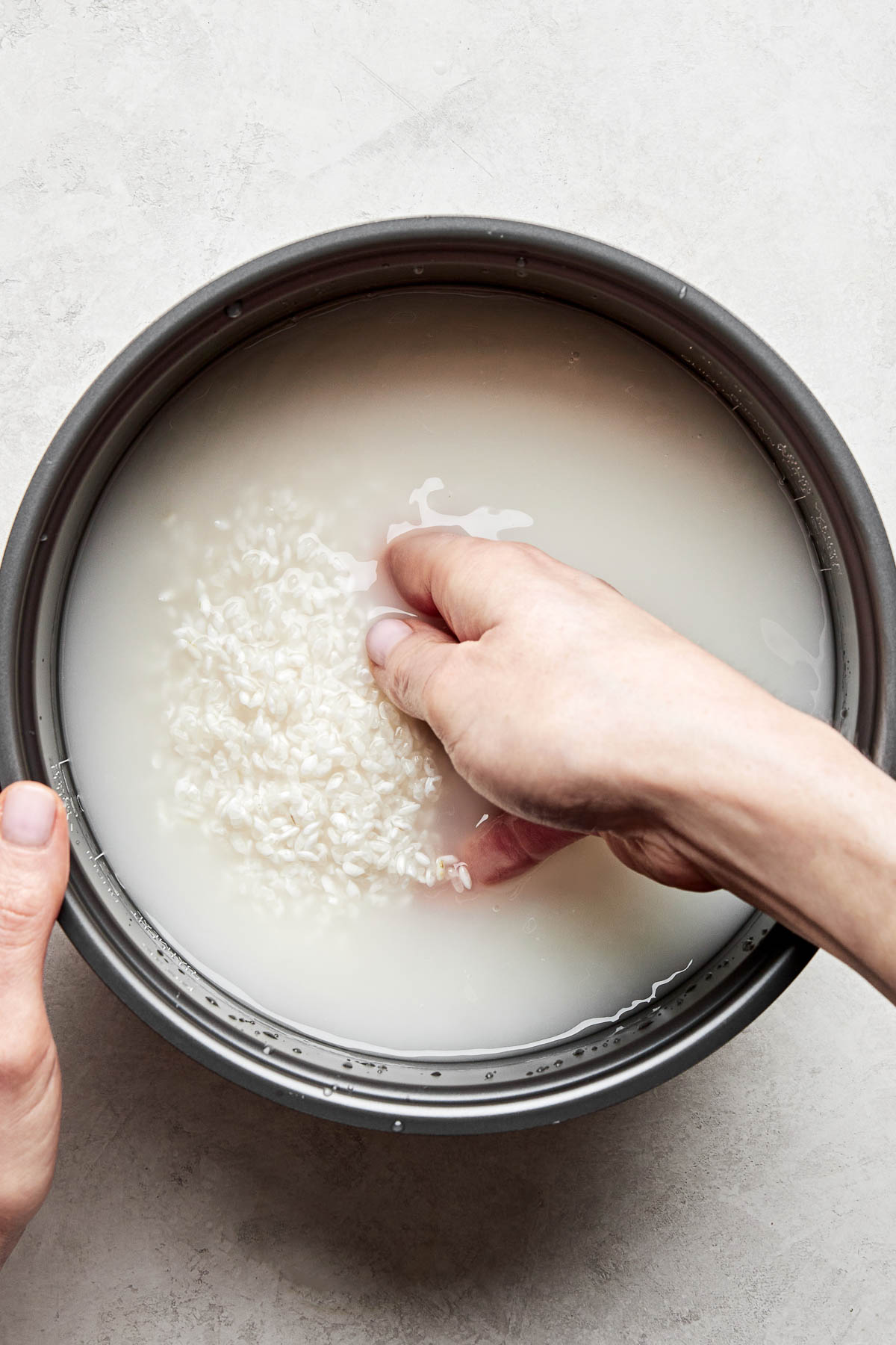 An overhead shot of a woman's hand rinsing rice in the bowl of a rice cooker atop a white surface.