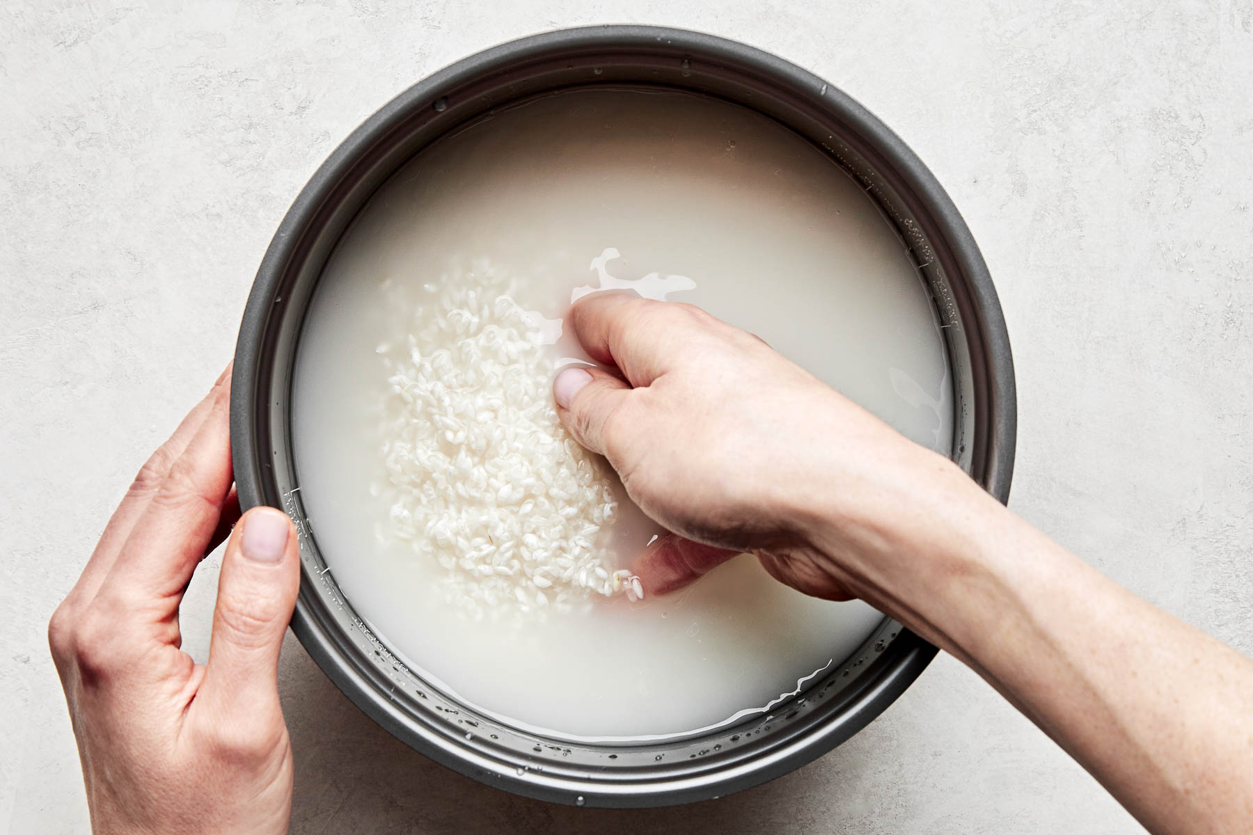 An overhead shot of a woman's hand rinsing rice in the bowl of a rice cooker atop a white surface.