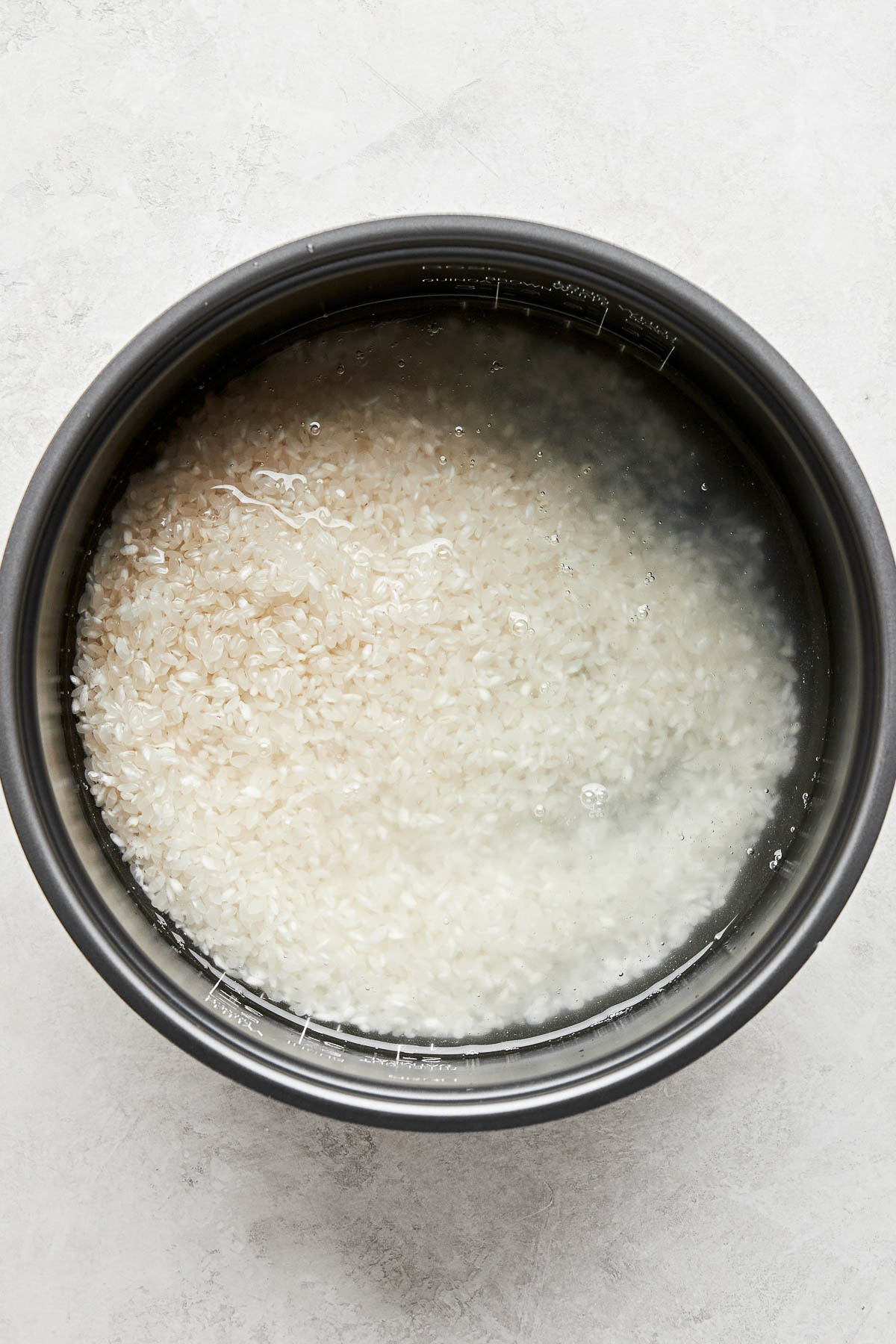 An overhead shot of uncooked rice and water in the bowl of a rice cooker atop a white surface.