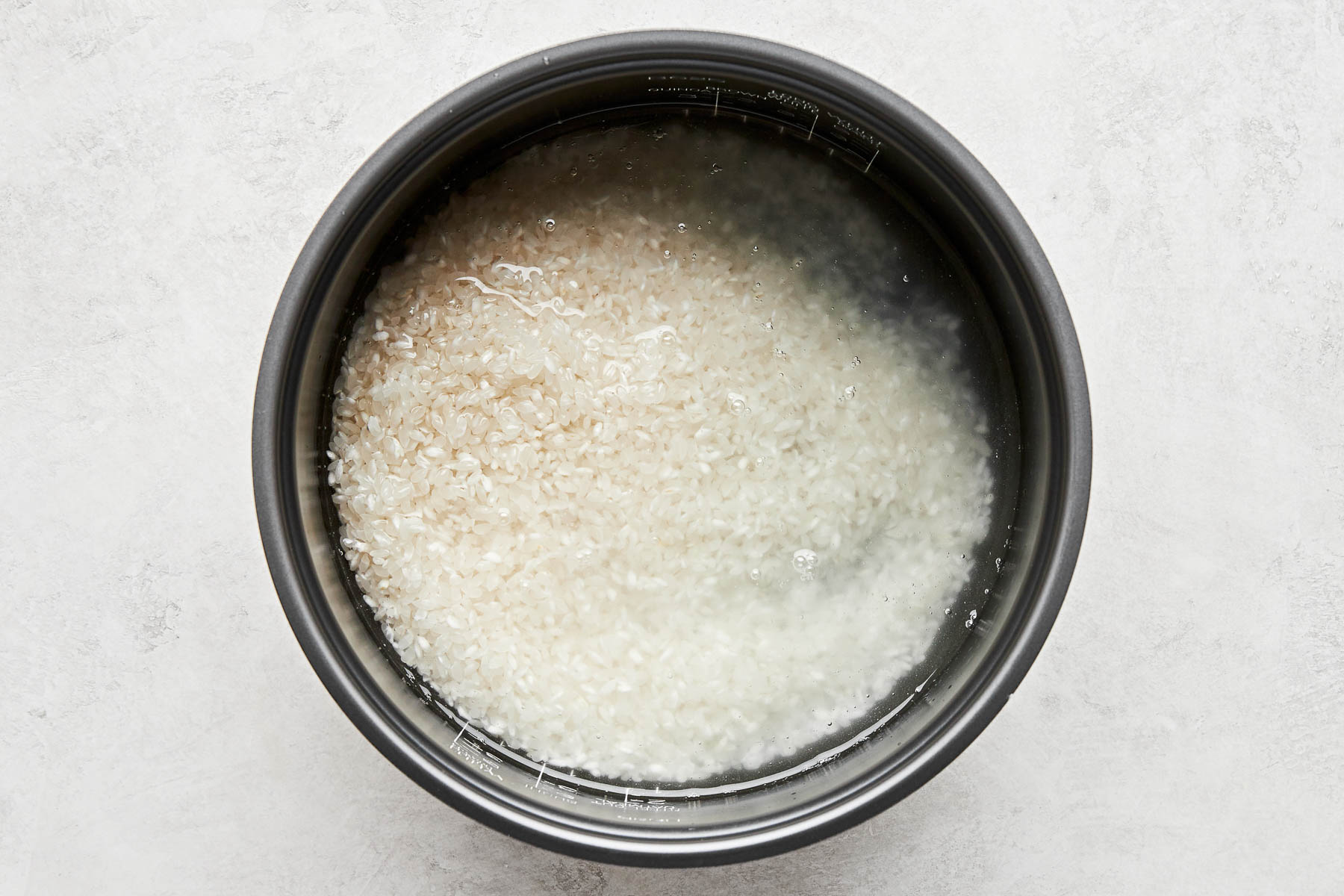 An overhead shot of uncooked rice and water in the bowl of a rice cooker atop a white surface.
