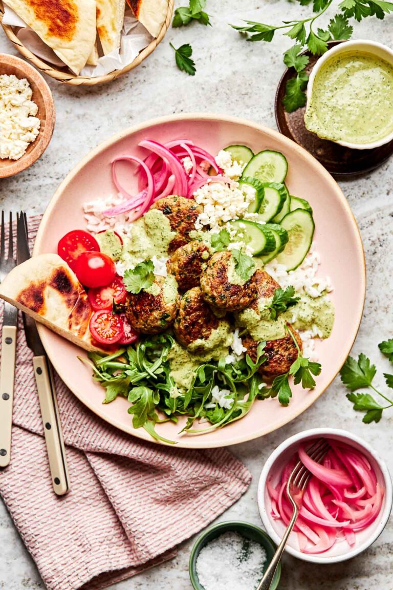 An overhead shot of falafel meatballs in a pink bowl with arugula, rice, halved tomatoes, tahini sauce, feta, cucumbers, pickled red onions, and a piece of pita bread. Bowls of pickled onions, tahini sauce, feta, salt and pita sit alongside it on a white textured surface. A pink cloth and a fork and knife sit next to the bowl.