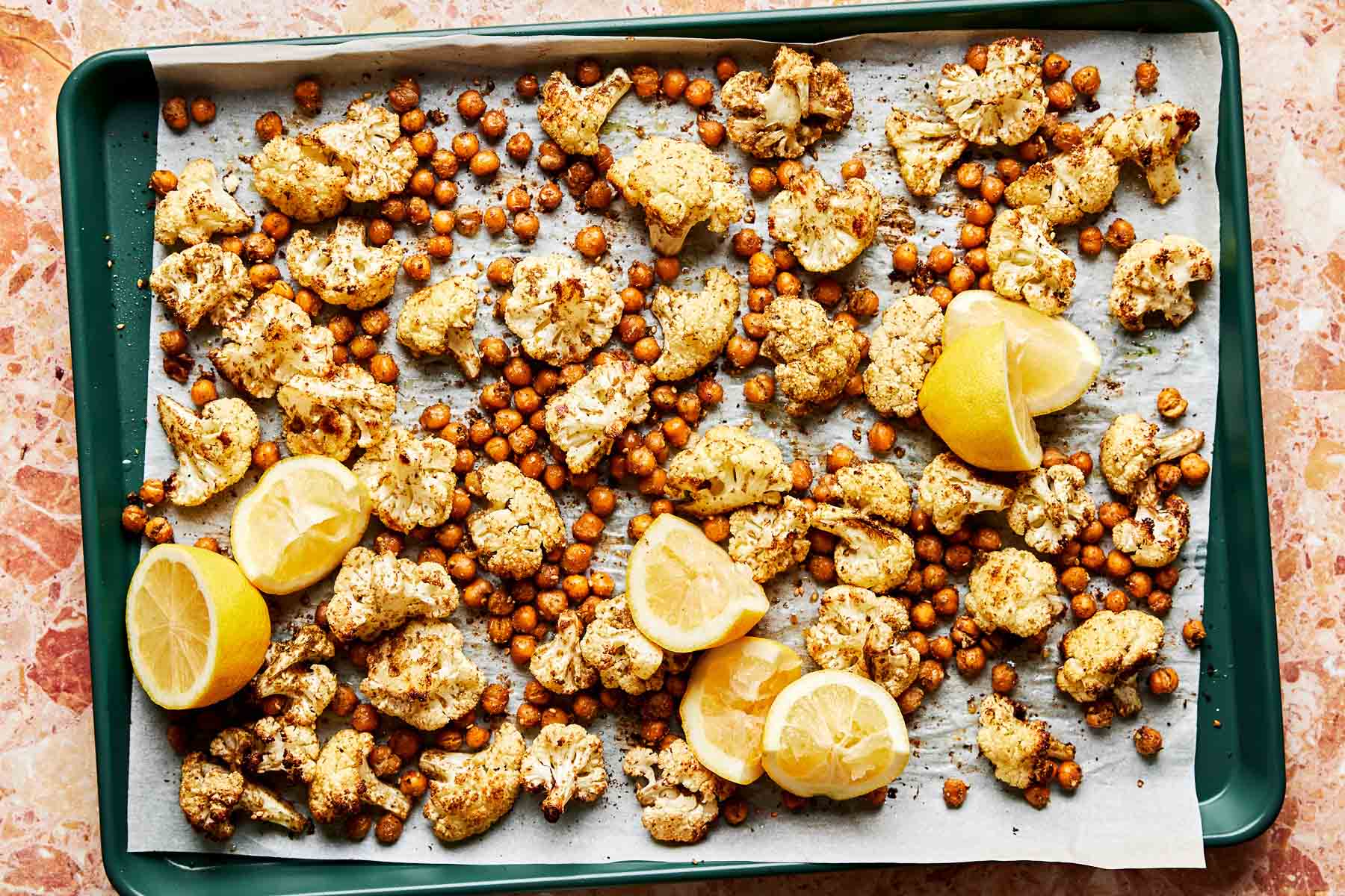 An overhead shot of roasted cauliflower and chickpeas with several lemon halves and wedges on a parchment-lined dark green baking sheet atop a light pink textured surface.