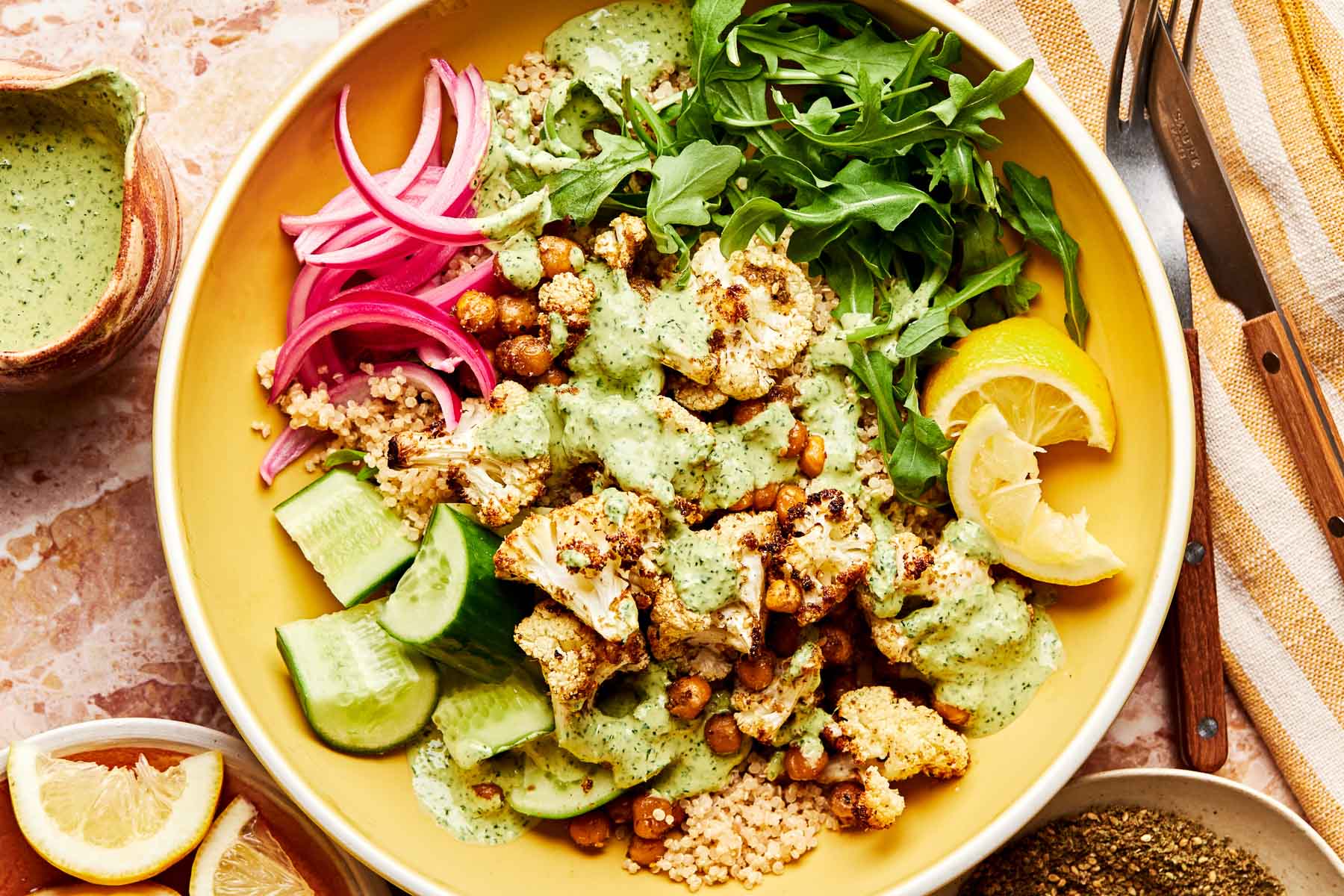 An overhead shot of quinoa, arugula, pickled red onions, cucumber slices, roasted cauliflower and chickpeas, tahini sauce and lemon wedges in a shallow yellow bowl atop a light pink surface. The bowl is surrounded with a striped cloth and small bowls and dishes of pepper, lemon wedges, and tahini sauce.