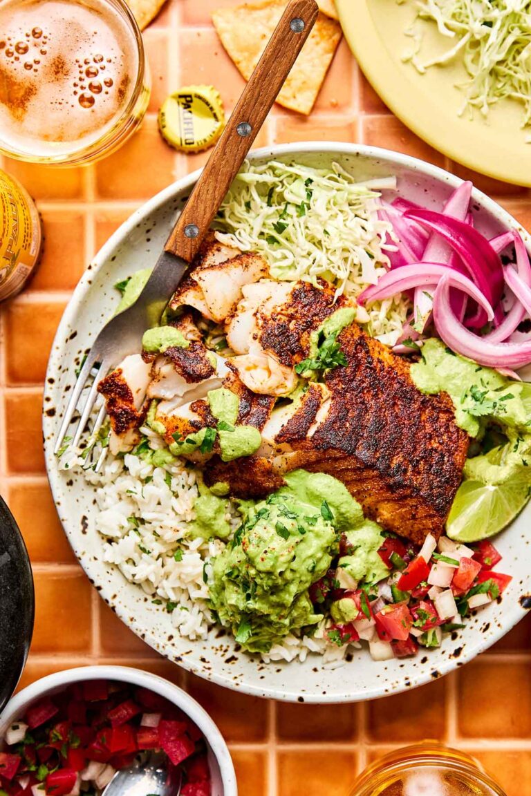 An overhead shot of a fish taco bowl with seasoned cod, rice, cabbage, red onion, guacamole, pico de gallo, and poblano ranch in a shallow stoneware bowl atop an orange tiled surface. Bowls of taco toppings sit alongside the bowl.