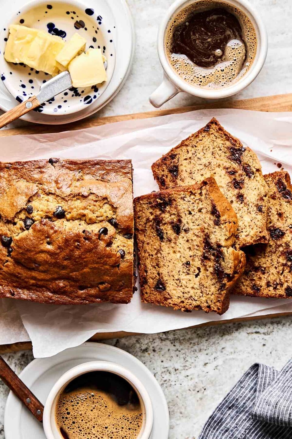 An overhead shot of 4 slices of chocolate chip banana bread and the remaining unsliced loaf on white parchment paper atop a wooden board. A dish of butter and two cups of coffee sit alongside the loaf of bread.