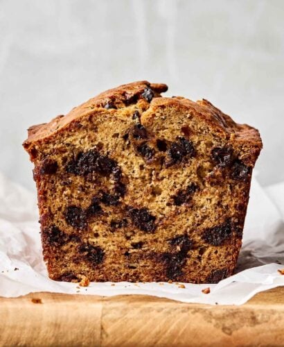 A close-up side shot of a halved loaf of banana bread, showing gooey chocolate chips. The bread sits on a parchment-lined wooden board atop a grey textured surface.