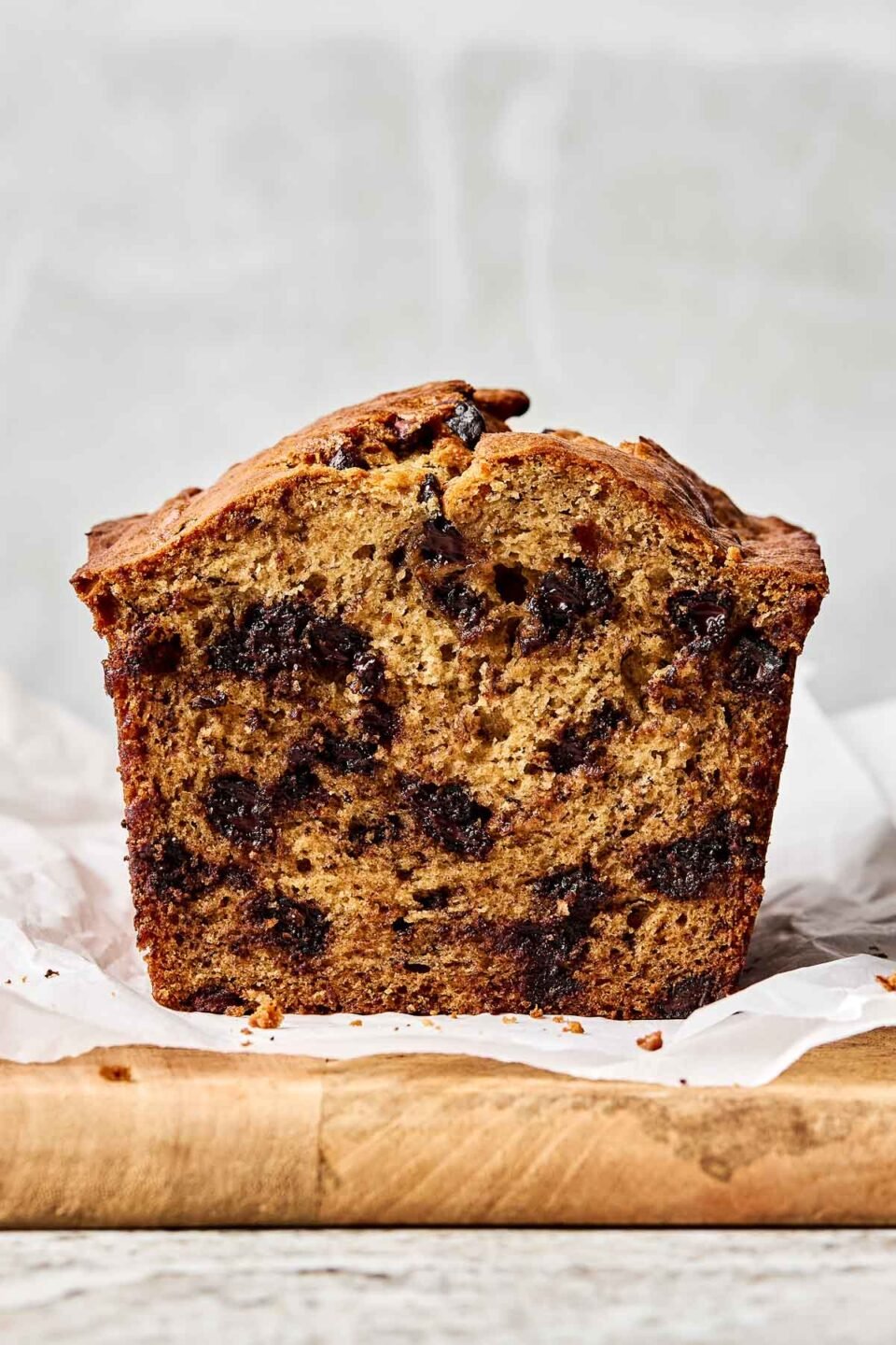 A close-up side shot of a halved loaf of banana bread, showing gooey chocolate chips. The bread sits on a parchment-lined wooden board atop a grey textured surface.