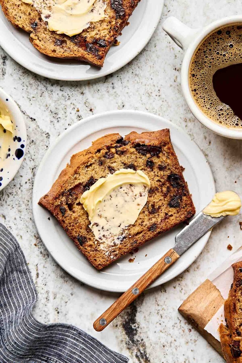 An overhead shot of a slice of chocolate chip banana bread slathered with butter on a white plate atop a grey textured surface. A butter knife with a pat of butter sits alongside it, and a second plate of banana bread, a cup of coffee, and the loaf of banana bread sit alongside the plate.